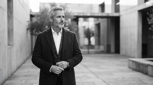 Cinematic black and white toned warm monochrome portrait of a mature male executive in his late 40s looking outward thoughtfully, soft woolen overcoat, outdoor architectural minimalist courtyard background with soft diffused morning light. Highly composed and resilient atmosphere.