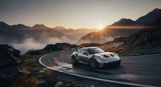 A pristine silver Porsche 911 GT3 RS speeding through winding misty alpine switchbacks in the Austrian Alps during golden dawn, captured with high-speed cinematic tracking, rich anamorphic lens flares, wet asphalt reflections, deep obsidian tones and amber sunrise glow
