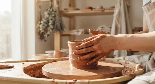 Sensory close-up of a potter shaping wet wild terracotta clay on a slow spinning wooden wheel in an airy, sun-drenched sunlit Japanese-inspired workshop atelier, dust particles floating in light.