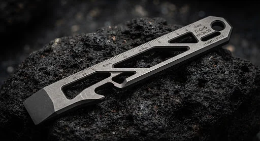 Skeletonized titanium Grade 5 pry bar tool with laser calibrated metric scale markings and integrated bottle lifter resting on charcoal volcanic stone. Clean industrial technical product shot with harsh directional light showcasing chamfered edges.