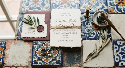 Refined detail still life of artisanal mexican wedding stationery. Hand-pressed deckled cotton paper, handwritten calligraphy in espresso ink, wax seal with olive sprig, antique gold rings on hand-painted Talavera ceramic tiles.