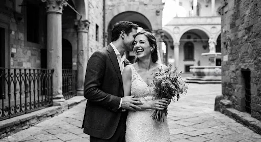 High-contrast black and white fine art photography of a candid romantic moment between lovers in a stone Renaissance courtyard in Florence Italy. The groom leans softly toward the bride's ear, genuine laughter, hands intertwined wearing vintage signet rings. Authentic Kodak Tri-X 400 deep blacks, luminous whites, cinematic grain texture.
