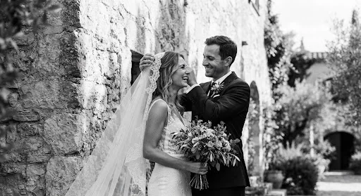Candid moment of groom gently fixing bride's veil against a sunlit stone wall in ancient Mediterranean estate, atmospheric shadows, editorial composition, black and white fine art print aesthetics
