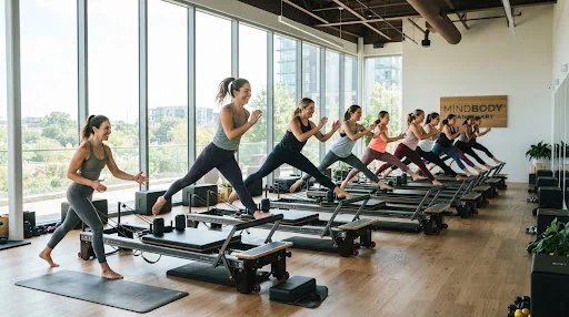 An energetic Reformer studio class showing padded jumpboards attached to machines, studio athletes working out in graceful synchrony with dramatic architectural daylight through floor-to-ceiling glass.